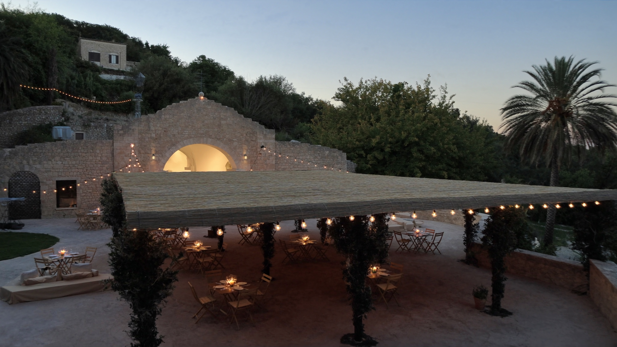 Terrazza di una masseria in pietra al crepuscolo, con tavolini sotto un pergolato illuminato da lucine